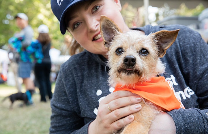 Lauren Ash wearing a long-sleeved Best Friends shirt and holding a small fluffy dog who is wearing an orange bandanna