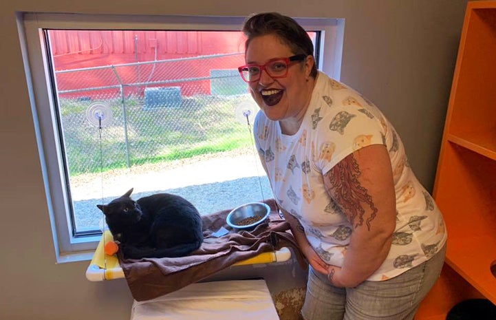 A smiling woman, Treah-Caldwell, next to a black cat next to a window