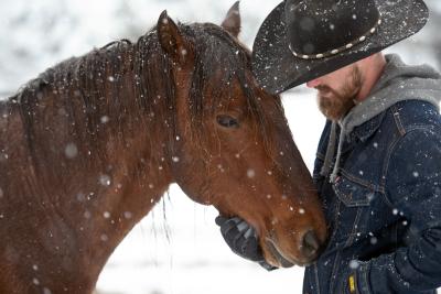 Christian Mathews wearing a hat and jacket affectionately holding the face of Byron the mustang while outside in the snow