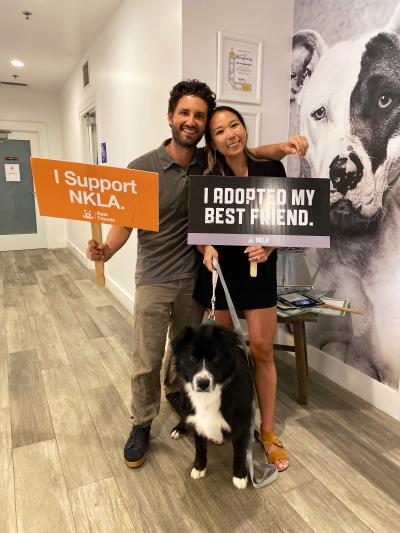 Couple holding two signs and standing with a black and white dog at the National Adoption Weekend