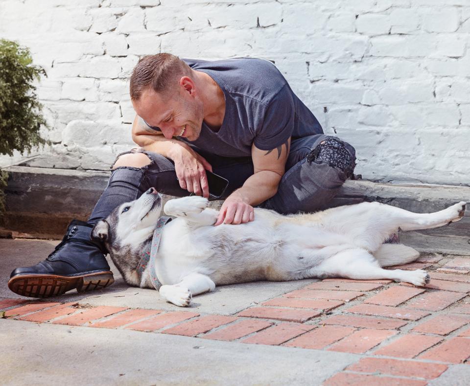 Seated smiling person petting a dog who is laying down outside on a stone patio