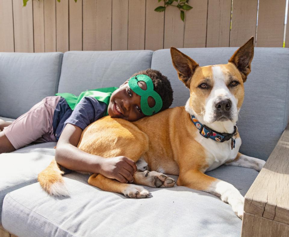 Child wearing a green superhero mask and cap lying on the back of a dog on a couch