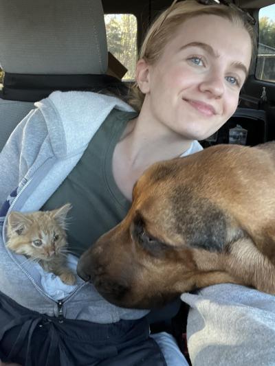 Emily Hirtle holding an orange kitten with a large brown dog's head in front of her