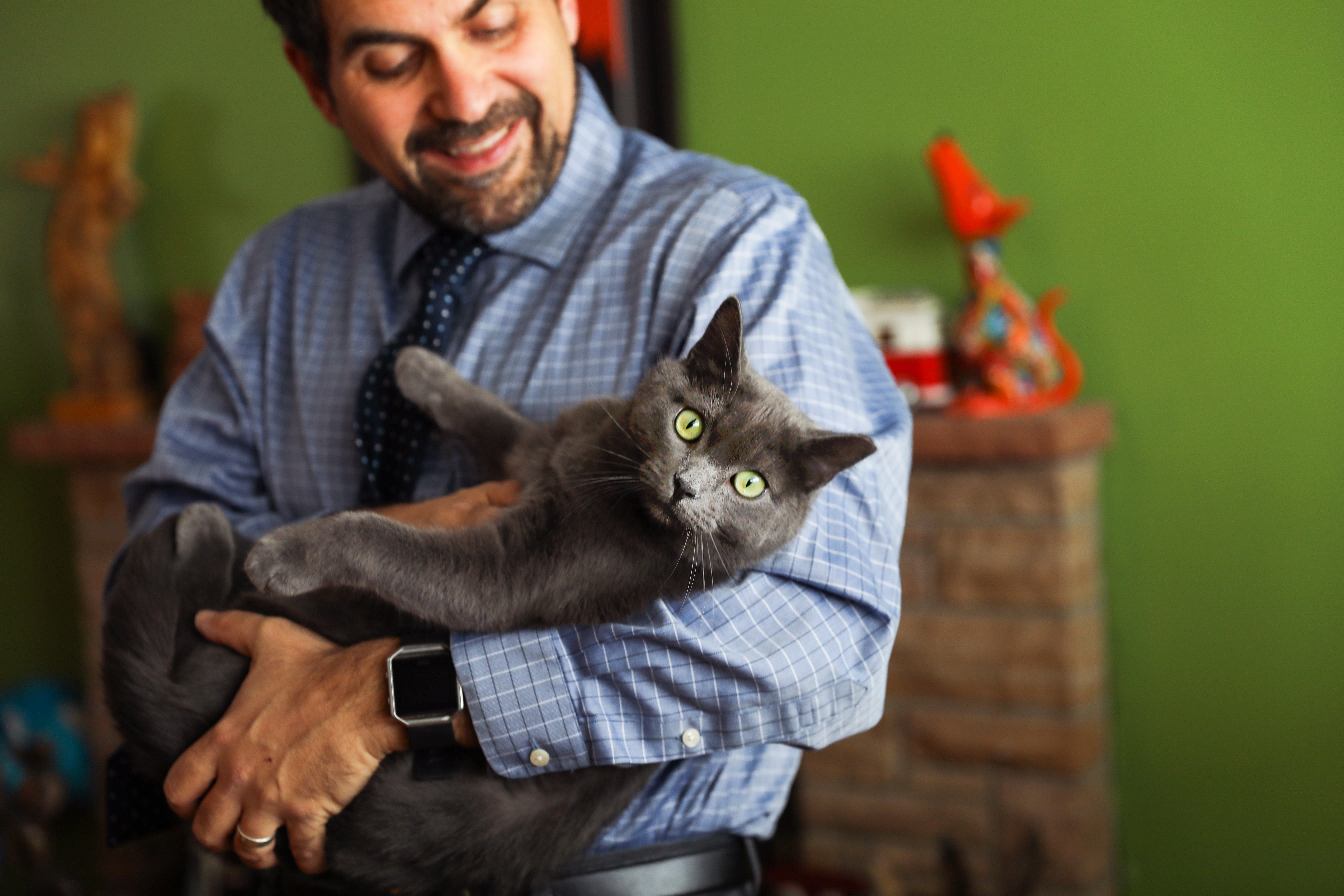 Smiling man cradling a gray cat in his arms like a baby, in front of a fireplace mantel