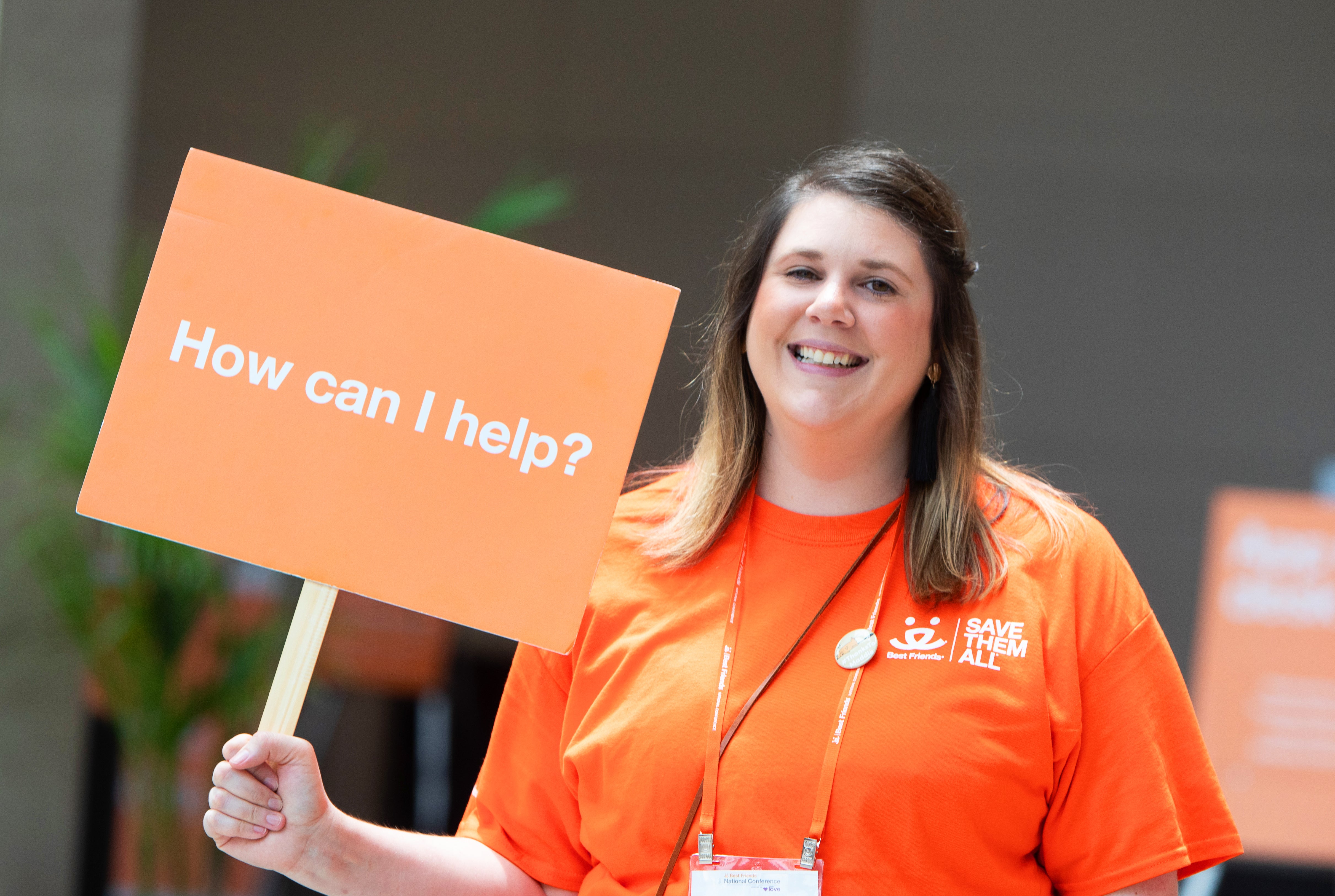 Smiling volunteer holding a "How can I help?" sign