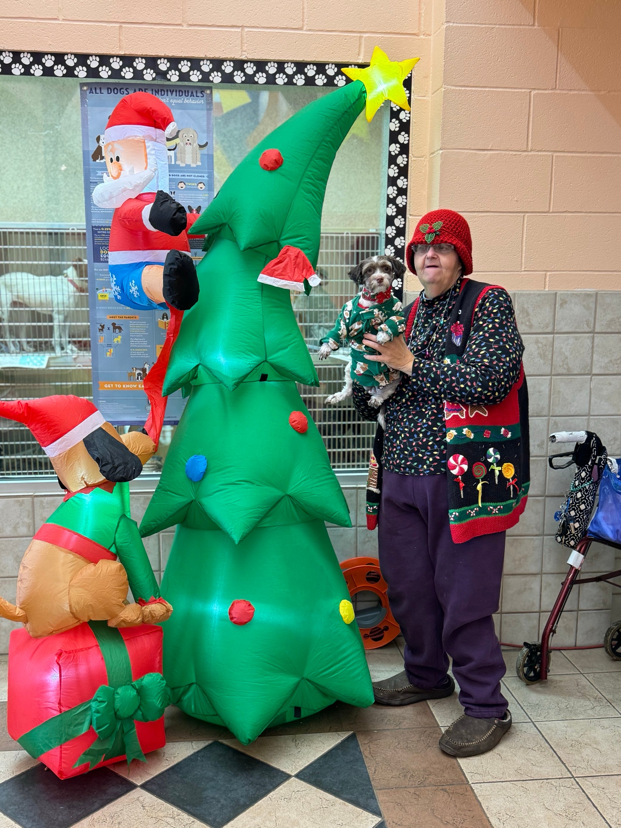 Persona con abrigo navideño con un perro pequeño con traje de navidad, al lado de un árbol inflado