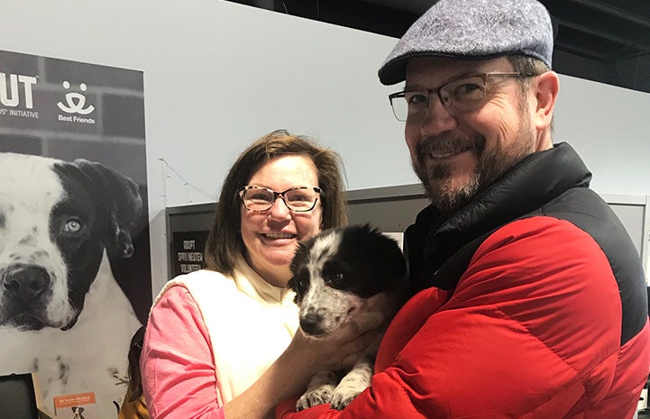 Man wearing a red coat and cap holding a black and white puppy and standing next to a smiling woman