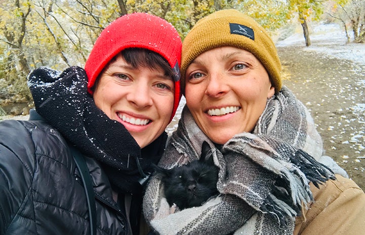 Two smiling women wearing hats and coats covered in a dusting of snow with Rafa the cat snuggled up in a scarf between them