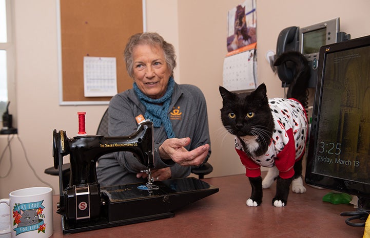 Gloria sitting next to a sewing machine behind Hero the cat wearing a red, black and white outfit