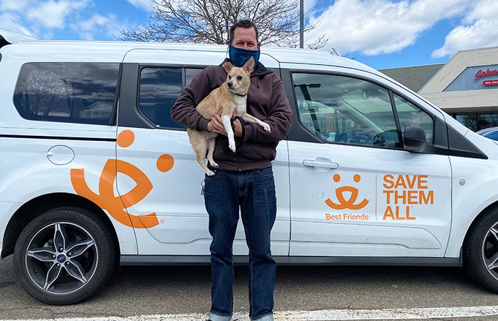 Richie the senior brown Chihuahua mix being held by a man wearing a mask standing in front of a Best Friends van