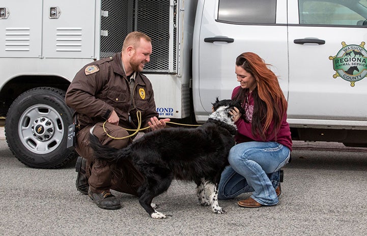 A male animal control officer kneeling down with a dog that a woman is hugging