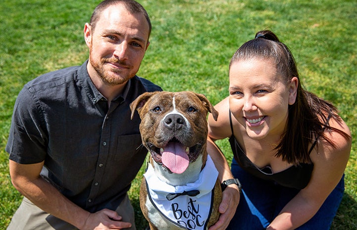 Smiling couple with a smiling dog wearing a bandanna that says "Best Dog" between them