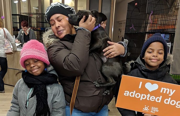 Woman carrying a black small black dog next to two children and one is holding a I heart my adopted dog sign