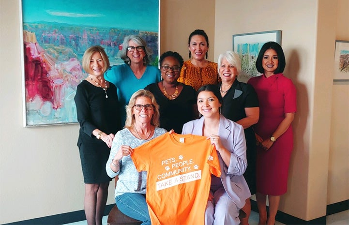Group of women standing in front of some paintings holding an orange T-shirt that says, "Pets, people, community, take a stand."