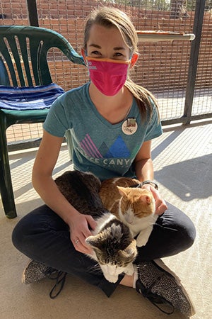 Tasha at Best Friends sitting on the ground inside a Cat World enclosure, with Drew and Jasper the cats lying on her lap