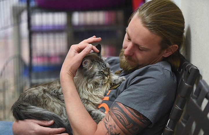 Caregiver Levi Myers sitting in a chair and cuddling Slader, a brown longhair tabby cat