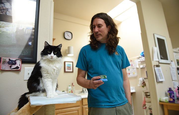 Zorro, the black and white tuxedo cat, sitting on a cat tree next to caregiver Eric, who is holding a can of food