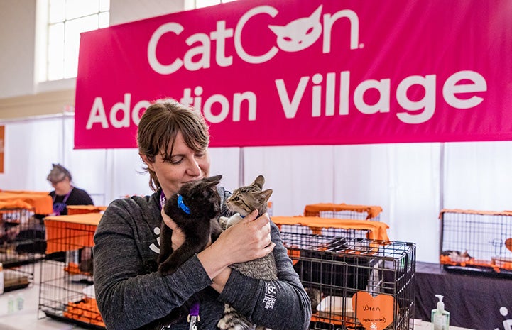 Woman holding two kittens in front of a CatCon Adoption Village sign