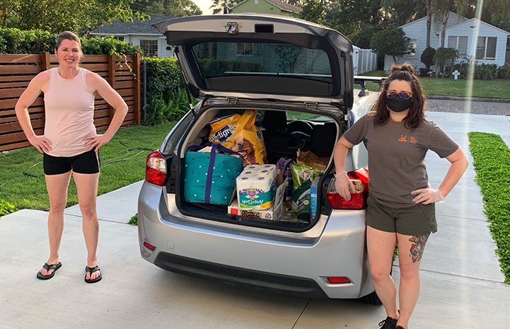 Two women standing next to an open trunk of a car holding supplies to be given to people in need