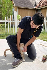 Man kneeling down to pet a cream and white cat with a few cans of cat food stacked next to them