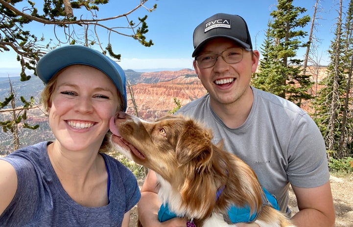 Dog sitting with two smiling people in front of red rock canyon view