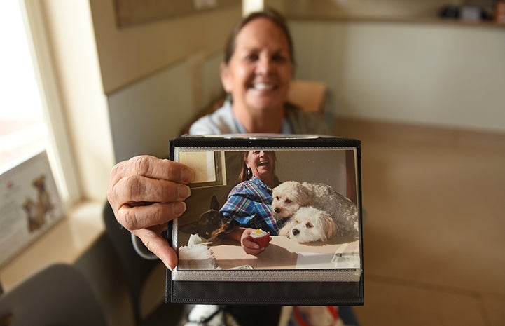 A smiling Pamela Zika holding a photo of her with Rascal and two other small white dogs