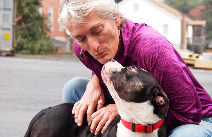 Woman in a purple jacket leaning in to kiss black and white dog