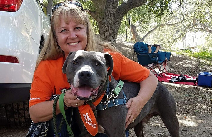 A woman wearing an orange T-shirt hugging a pit-bull-terrier-type dog while on a walk