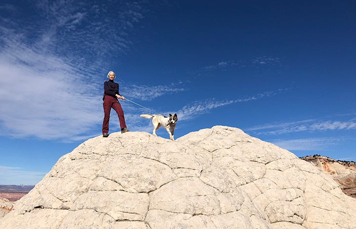 Volunteer Heather Harding hiking with Sun the dog on top of a beautiful rock formation with blue sky and clouds behind them