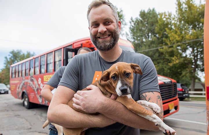 Smiling man standing in front of the transport bus holding a tri-colored puppy