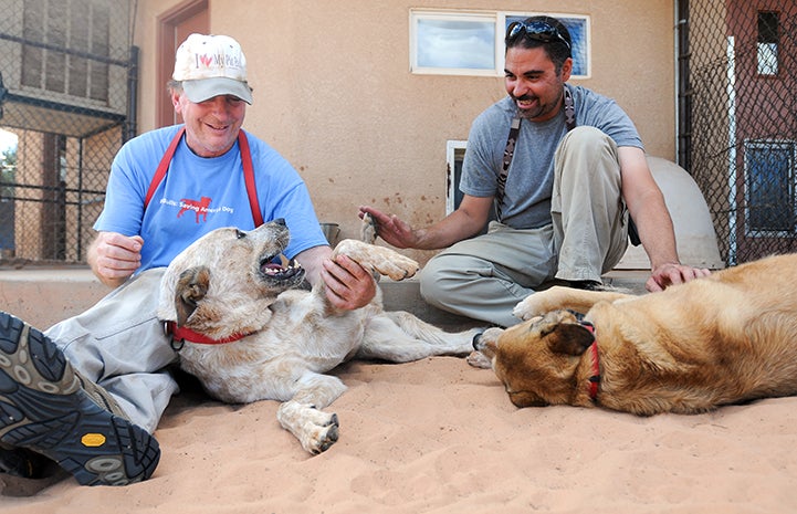 Two male caregivers, Tom and Haven, sitting on the ground petting two dogs, Fred and Express