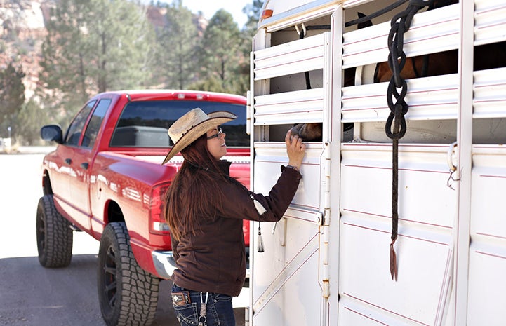 Jessica, wearing a cowboy hat, smiling and putting her hand into the trailer to pet a horse