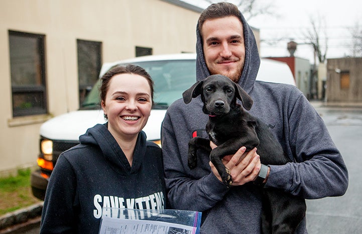 Young couple standing outside, smiling and holding a black puppy