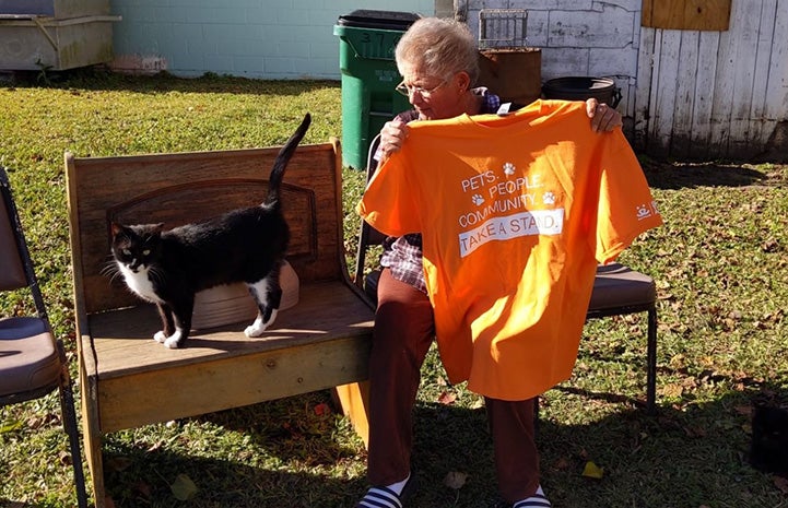 Person sitting down and holding an orange Best Friends Pets People Community Take a Stand T-shirt, next to a black and white cat on a wooden bench