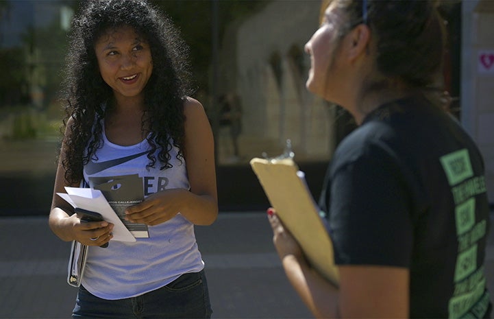 Person holding a clipboard talking to another person smiling and holding paperwork