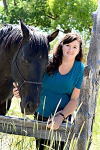 Smiling woman wearing a teal shirt leaning on a fence with a dark brown horse next to her