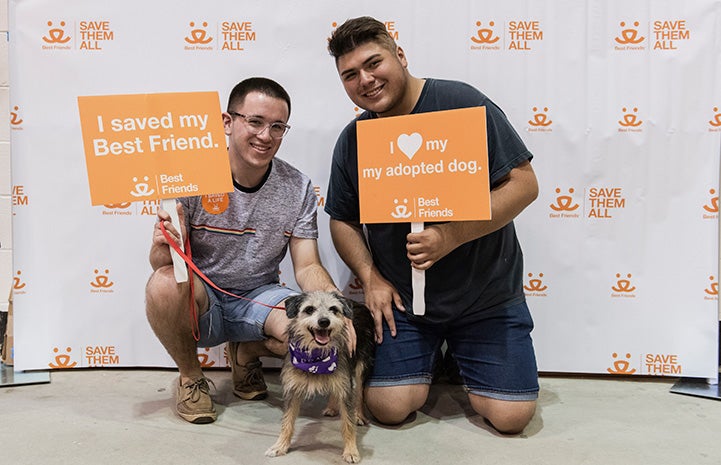 Two young men holding adoption signs in front of a Best Friends backdrop with the dog they'd just adopted at the Houston Super Adoption