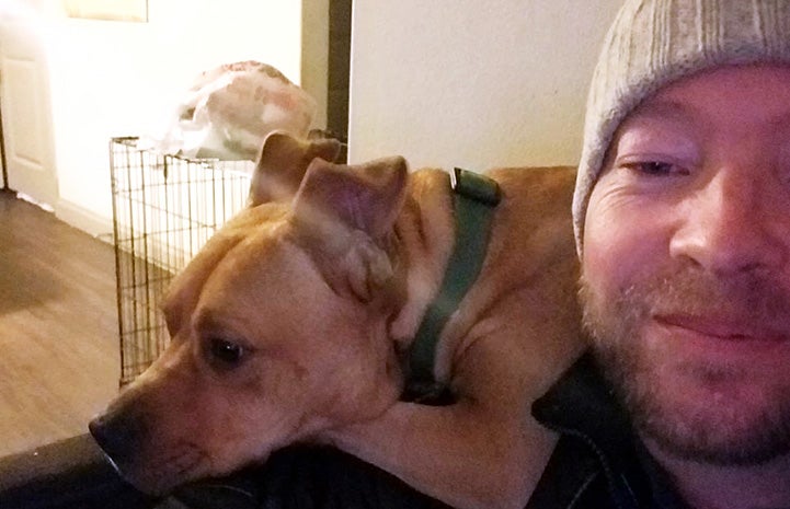 Samwell the dog lying behind a man who is wearing a knit cap's shoulder