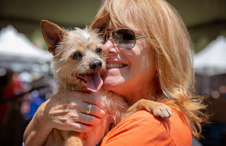 A smiling woman wearing sunglasses and an orange T-shirt, holding a fluffy terrier mix dog whose tongue is out and paw on her shoulder