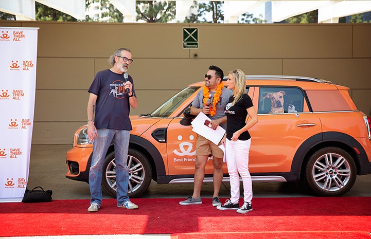 Three people talking in front of an orange Best Friends-branded MINI car at the A tent with human and dog activity at the NKLA Pet Super Adoption