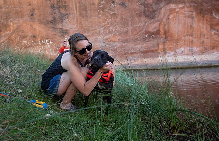 Person hugging a dog out on the grass with a red cliff behind them