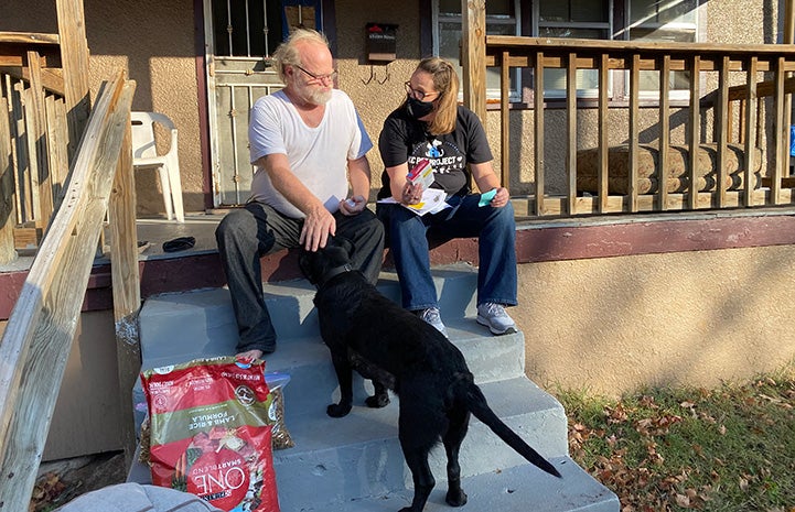 Kansas City Pet Project animal services team member on a porch giving a man some dog food while the man pets a black Lab