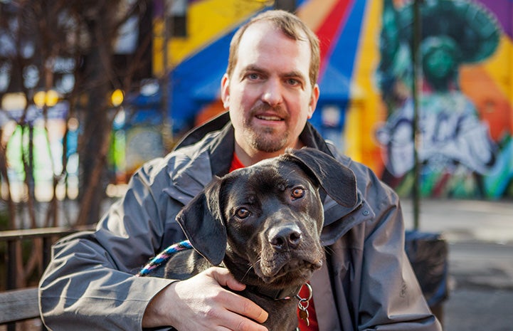 Volunteer Fabio Vitolla with his arm around a large black dog with a colorful background behind them
