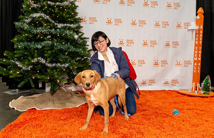 Woman and brown dog on an orange carpet in front of a Best Friends backdrop and Christmas tree