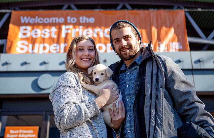 Couple holding a small blond puppy in front of a New York Super Adoption banner