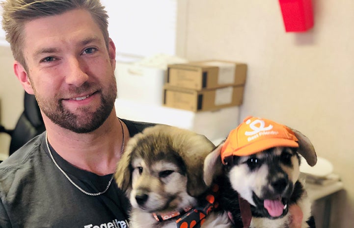 Man holding two adorable puppies, with one of the puppies wearing an orange Best Friends hat