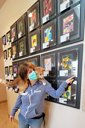 Woman with a cat on her shoulders pointing at the wall of cat paintings