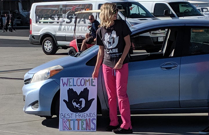 Woman at airport waiting to pick up transported animals with a sign that says, "Welcome Best Friends kittens"