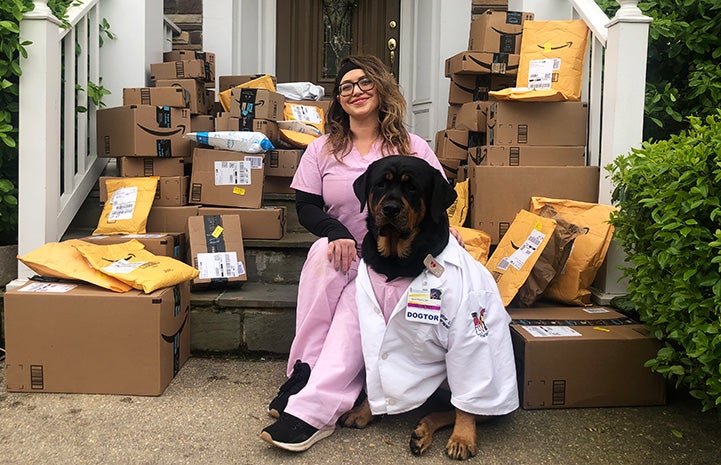 Caroline Benzel sitting on some steps with Loki the dog wearing a doctor's coat, surrounded by lots of mailed packages