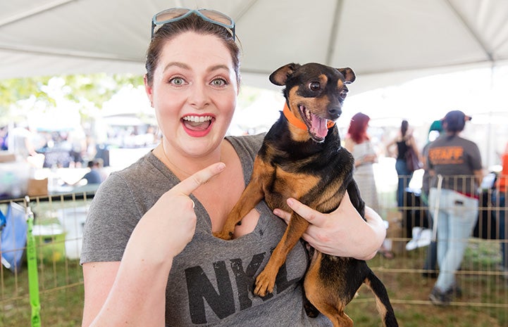 Smiling Lauren Ash wearing a NKLA T-shirt and pointing at a small black and brown dog who she's holding